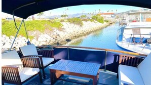Cubierta de barco con una mesa y sillas, vista de un canal con edificios y vegetación.