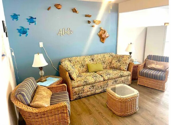 A living room with a floral-patterned sofa, two wicker chairs, a small table, and wall decorations including a 'Aloha' sign and turtle figurines.