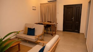 A living room with a sofa, a wooden table, and a chair. There is a door and an air conditioning unit on the wall. - Lokko Serene One Bedroom (Accra)