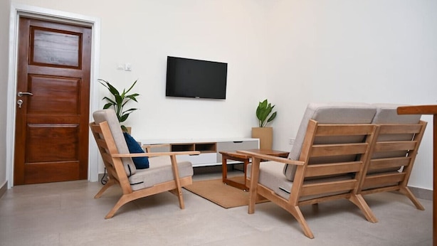 A living room with a wooden door, a flat-screen TV mounted on the wall, and two wooden chairs with cushions. - Lokko Serene One Bedroom (Accra)