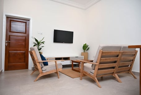 A living room with a wooden door, a flat-screen TV mounted on the wall, and two wooden chairs with cushions.