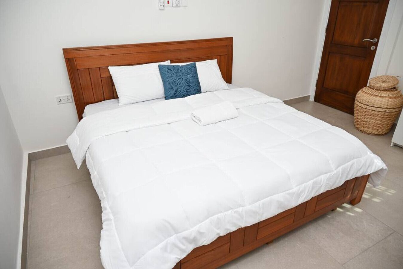 A neatly made bed with white bedding and a wooden headboard in a room with a tiled floor and a wooden door.