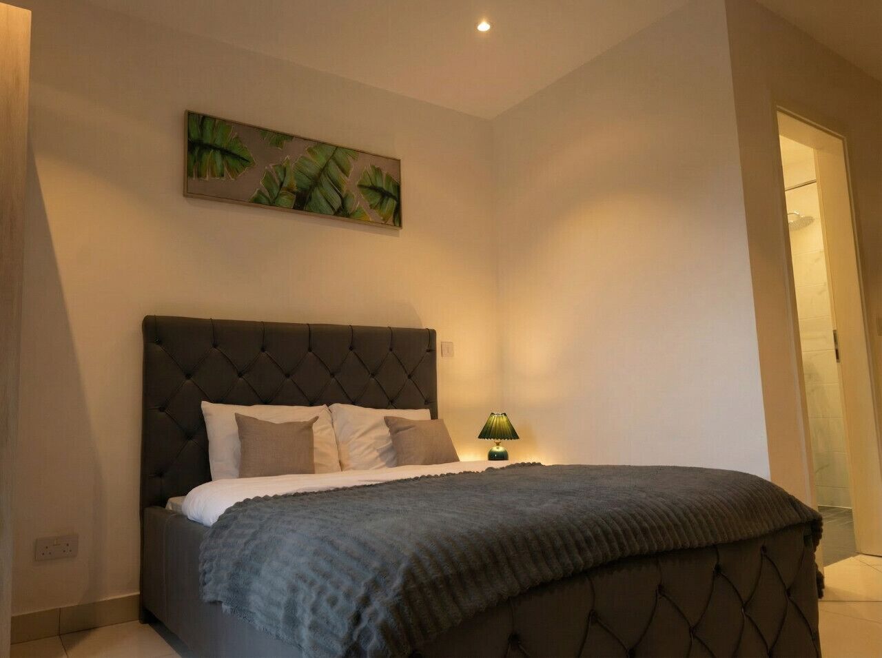 A bedroom with a tufted headboard, a bed with white and brown bedding, a green lamp on a bedside table, and a painting of leaves on the wall.