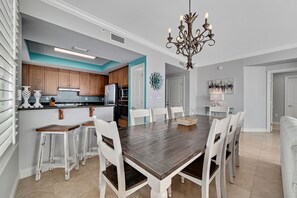 A dining area with a wooden table and white chairs, a kitchen with wooden cabinets, and a chandelier. - Indigo 1806E | Resort Amenities | Heated Pool | My Beach Getaways (Pensacola)