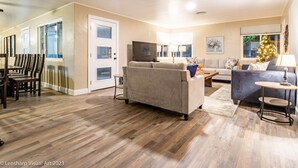 A spacious living room with a beige sofa, a blue armchair, and a coffee table. There is a dining area with a wooden table and chairs. A Christmas tree is visible in the corner. - Ultimate Relaxation Retreat w/ Game Room & Hot Tub (Ahwahnee)