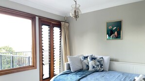 Une chambre avec une grande fenêtre, un lit recouvert d’une couverture bleue et un tableau accroché au mur.