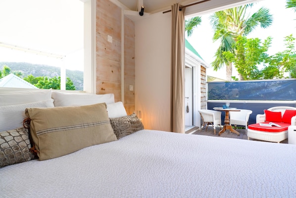 Une chambre dotée d'un grand lit, d'un balcon avec des meubles en osier blancs et offrant une vue sur la verdure.
