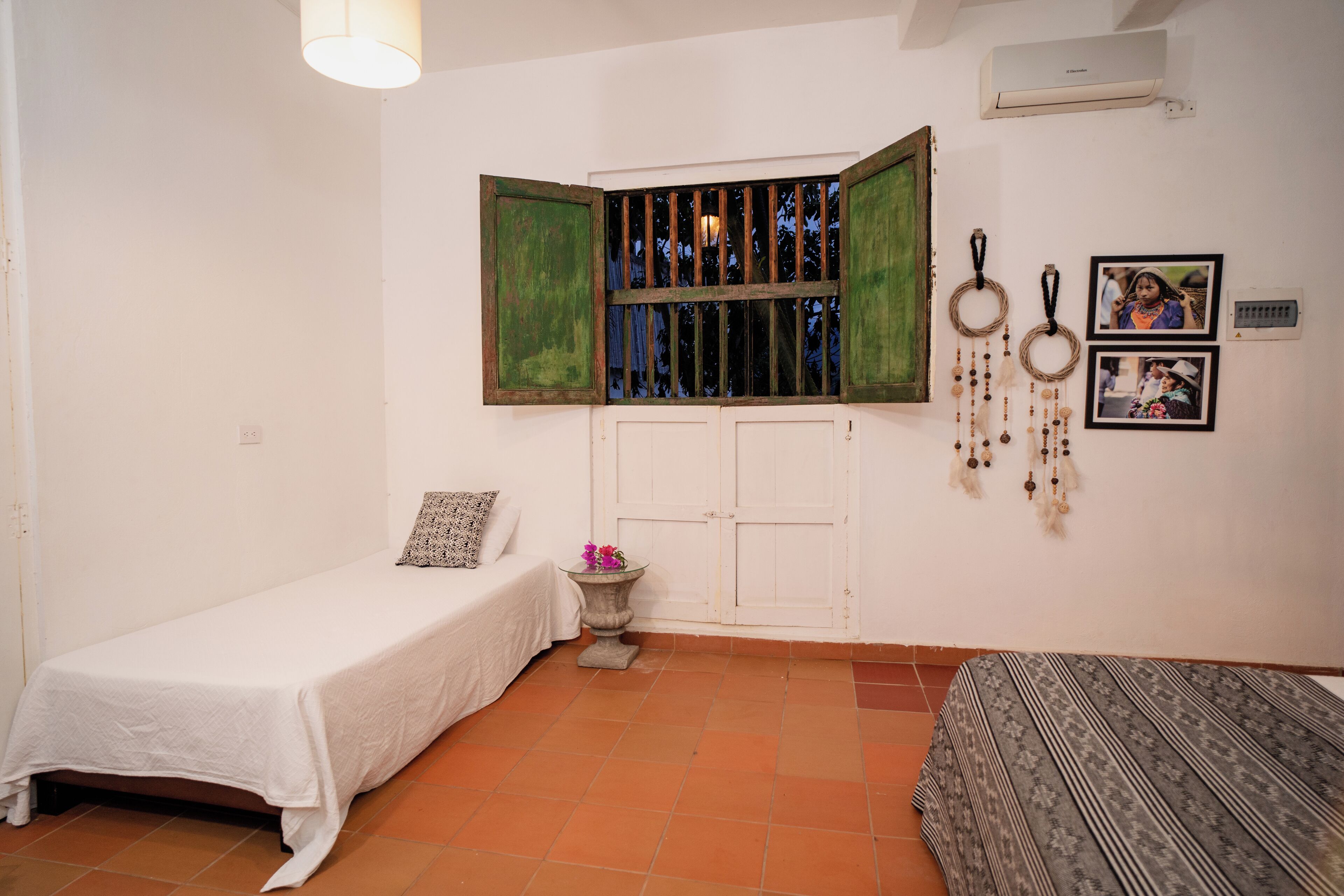 A small, clean room with a single bed, a small table with a vase, and a window with green shutters.