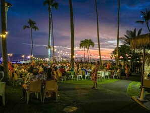 An outdoor dining area with tables and chairs, surrounded by palm trees, under a twilight sky.