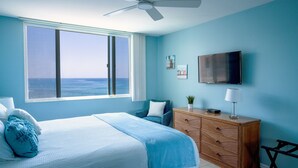 Un dormitorio con un ventanal grande, una cama con ropa de cama azul, un aparador de madera, un televisor en la pared y un ventilador de techo.