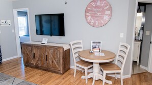 A dining area with a wooden table and white chairs, a large clock on the wall, and a TV mounted above the table.