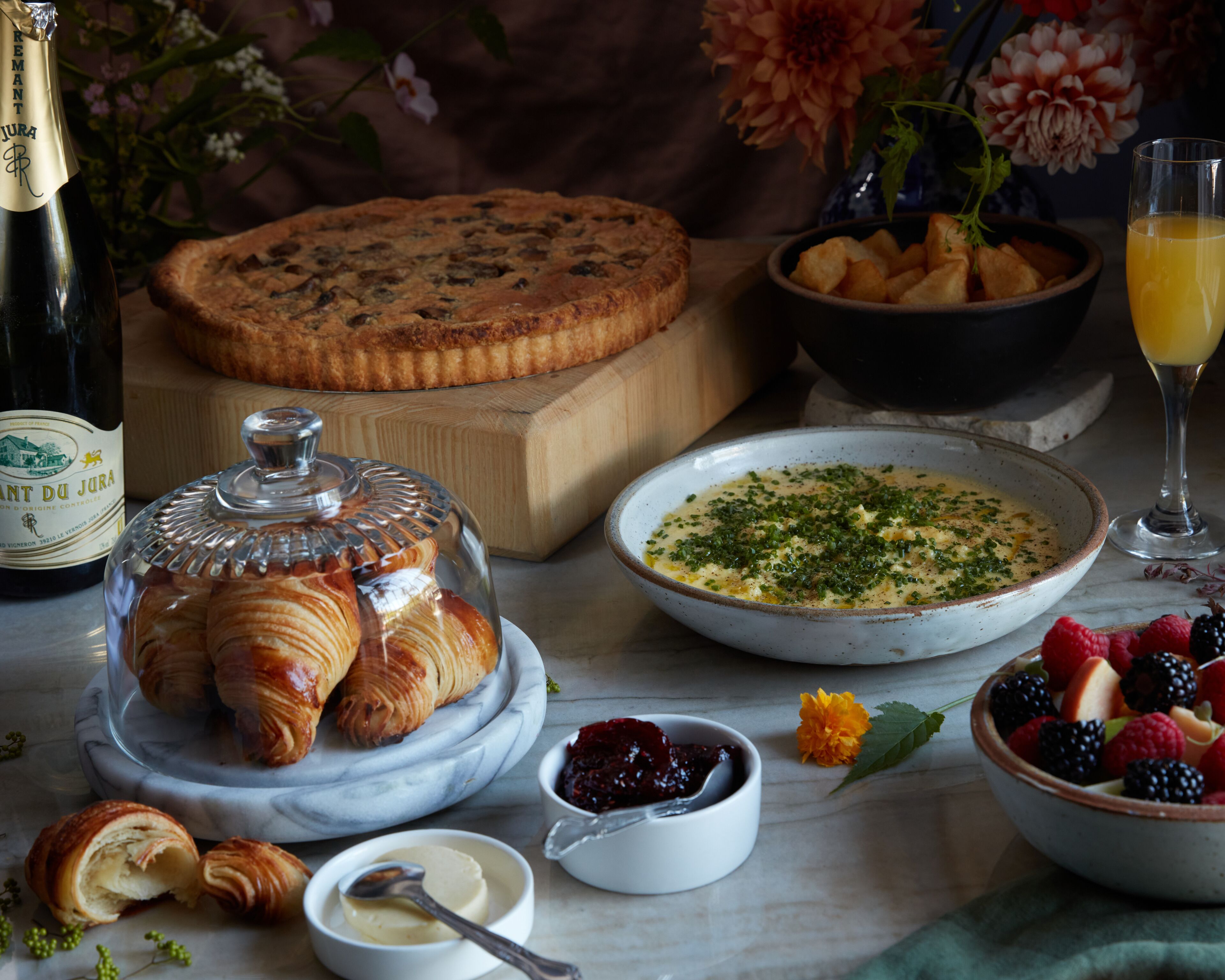 A table with croissants, a fruit bowl, a bowl of soup, a bottle of wine, and a glass of orange juice.