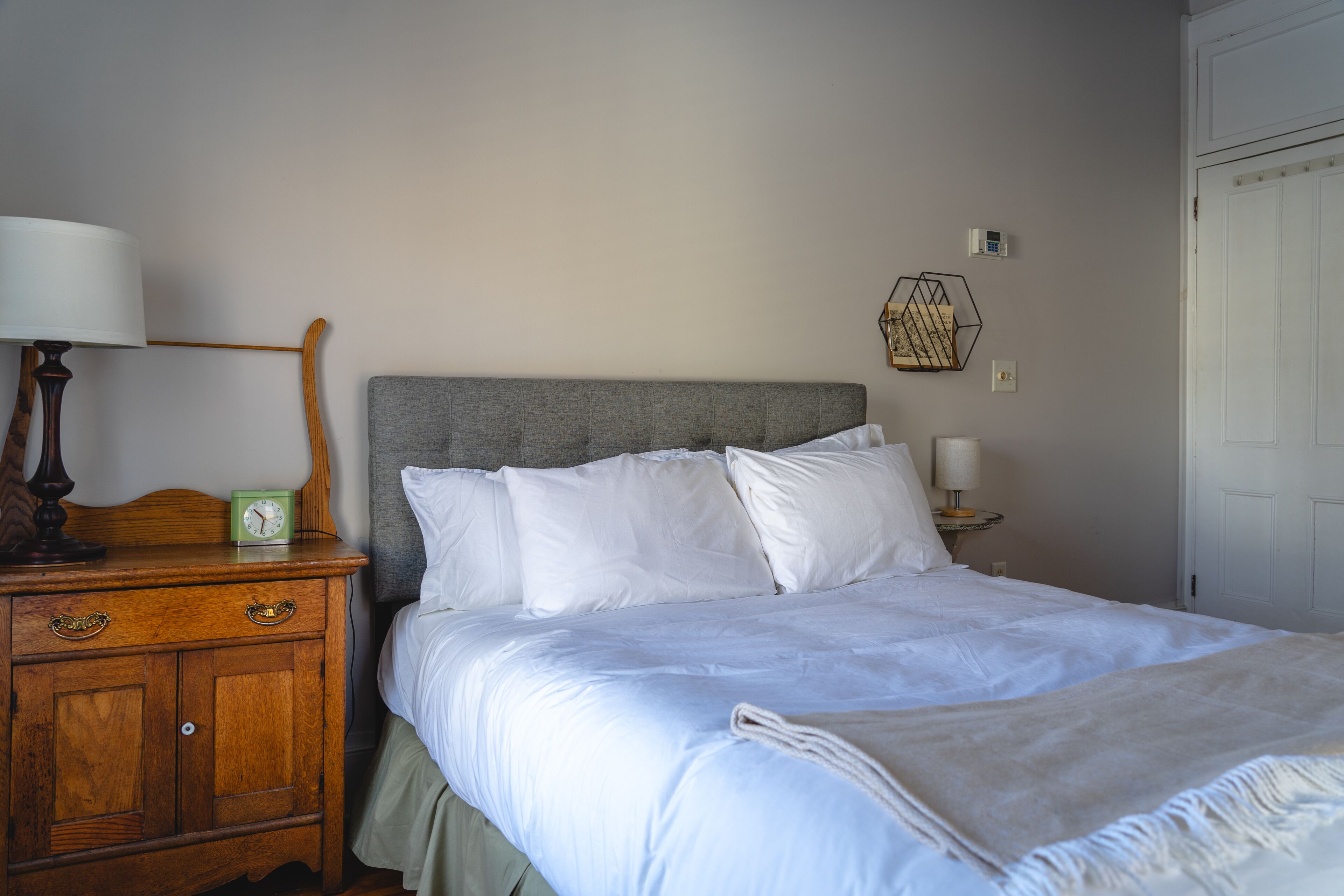 A neatly made bed with white linens, a wooden nightstand with a lamp and a clock, and a wall-mounted light fixture.