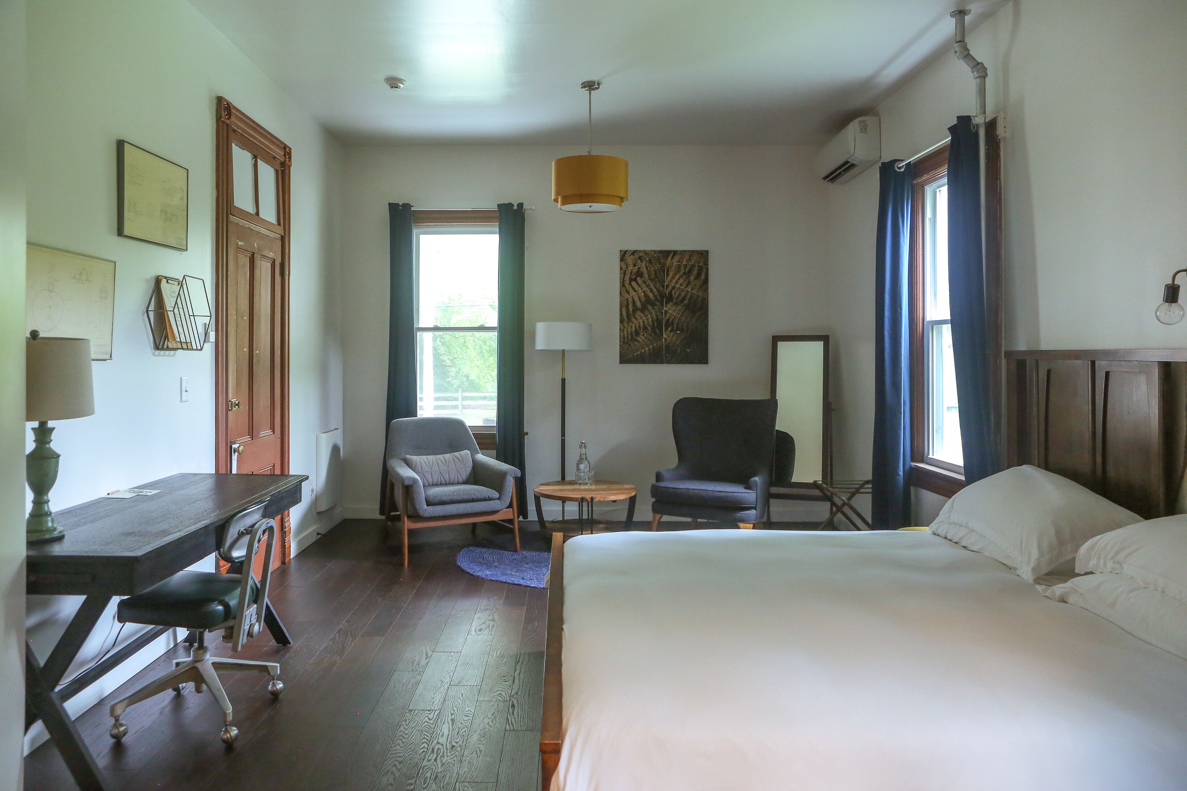 A hotel room with a bed, desk, two chairs, a small table, and a window with curtains.