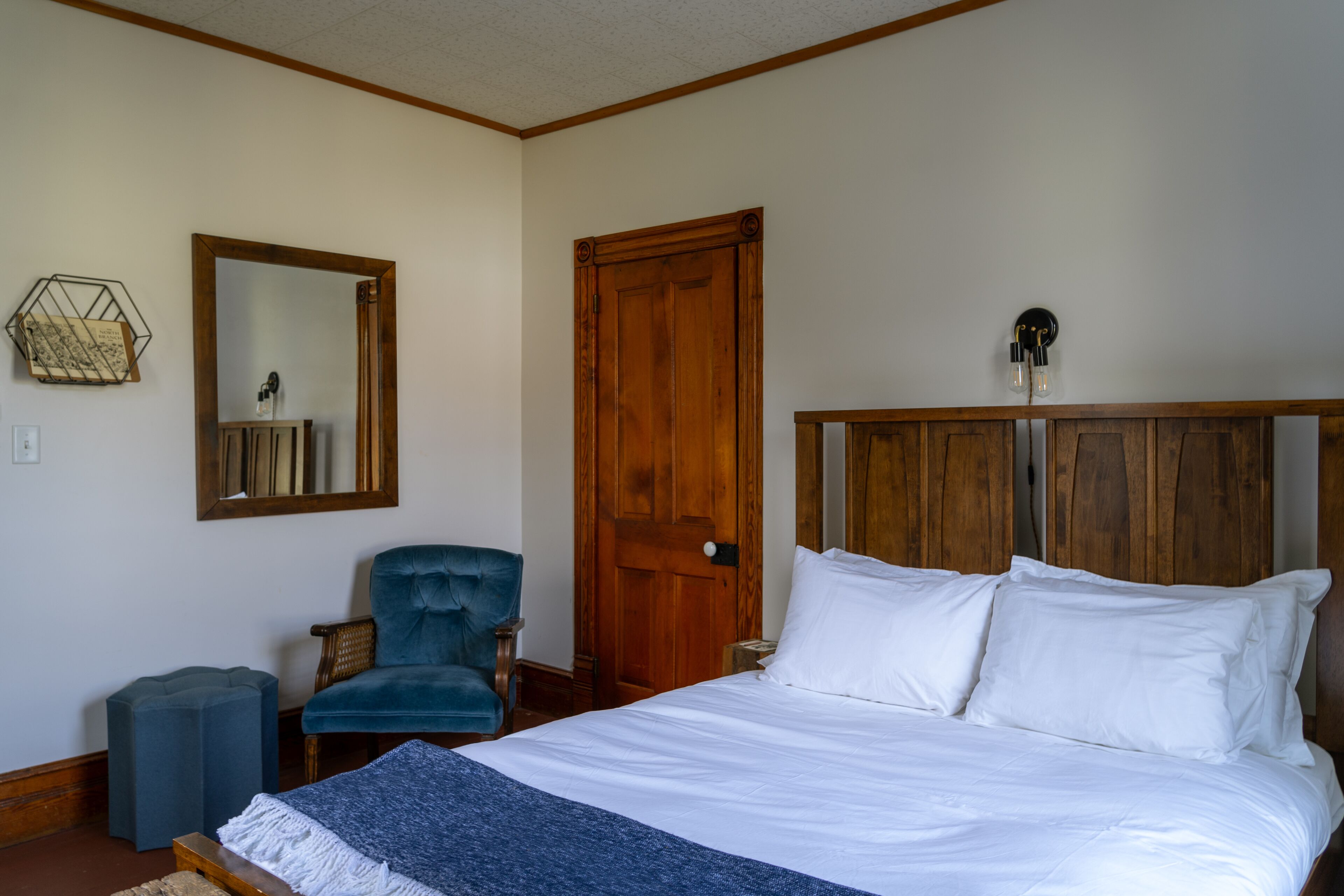 Un dormitorio con una cama grande, cabecera de madera, un sillón azul, un pequeño reposapiés y una puerta de madera.
