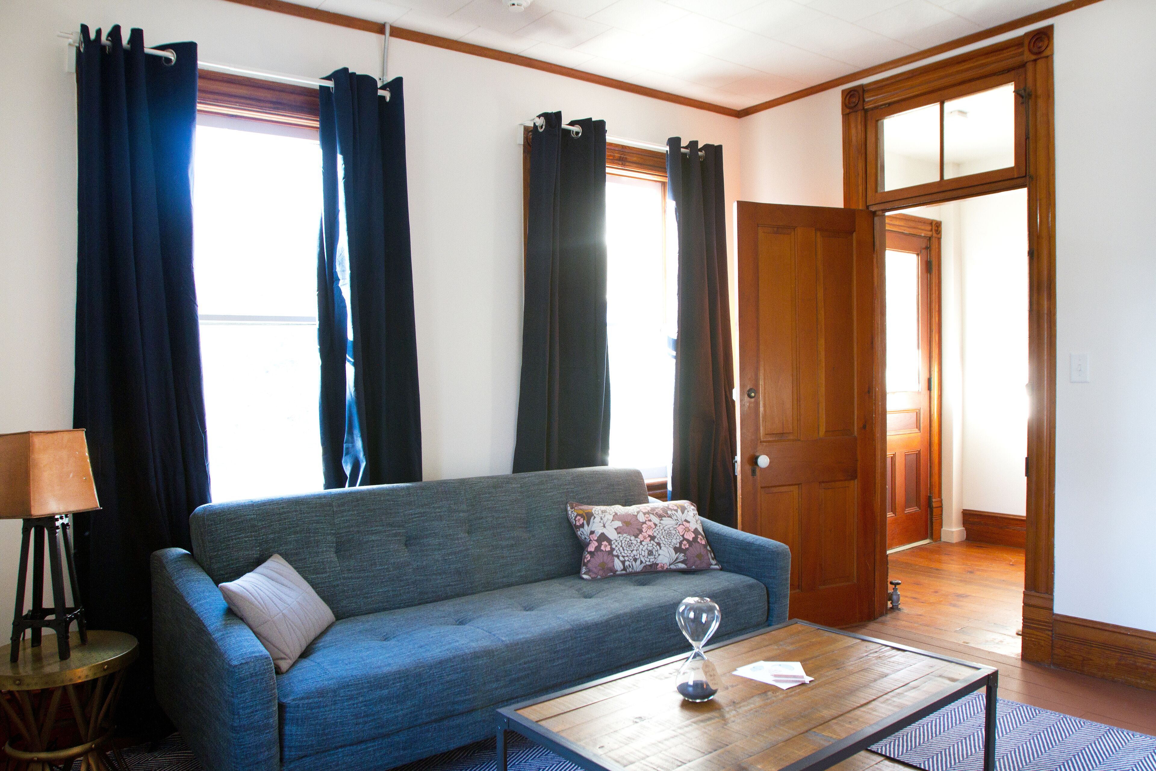 A living room with a sofa, a coffee table, and a lamp. There are curtains on the windows and a door leading to another room.