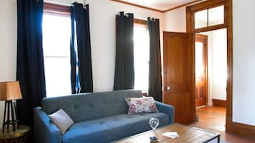 A living room with a sofa, a coffee table, and a lamp. There are curtains on the windows and a door leading to another room.