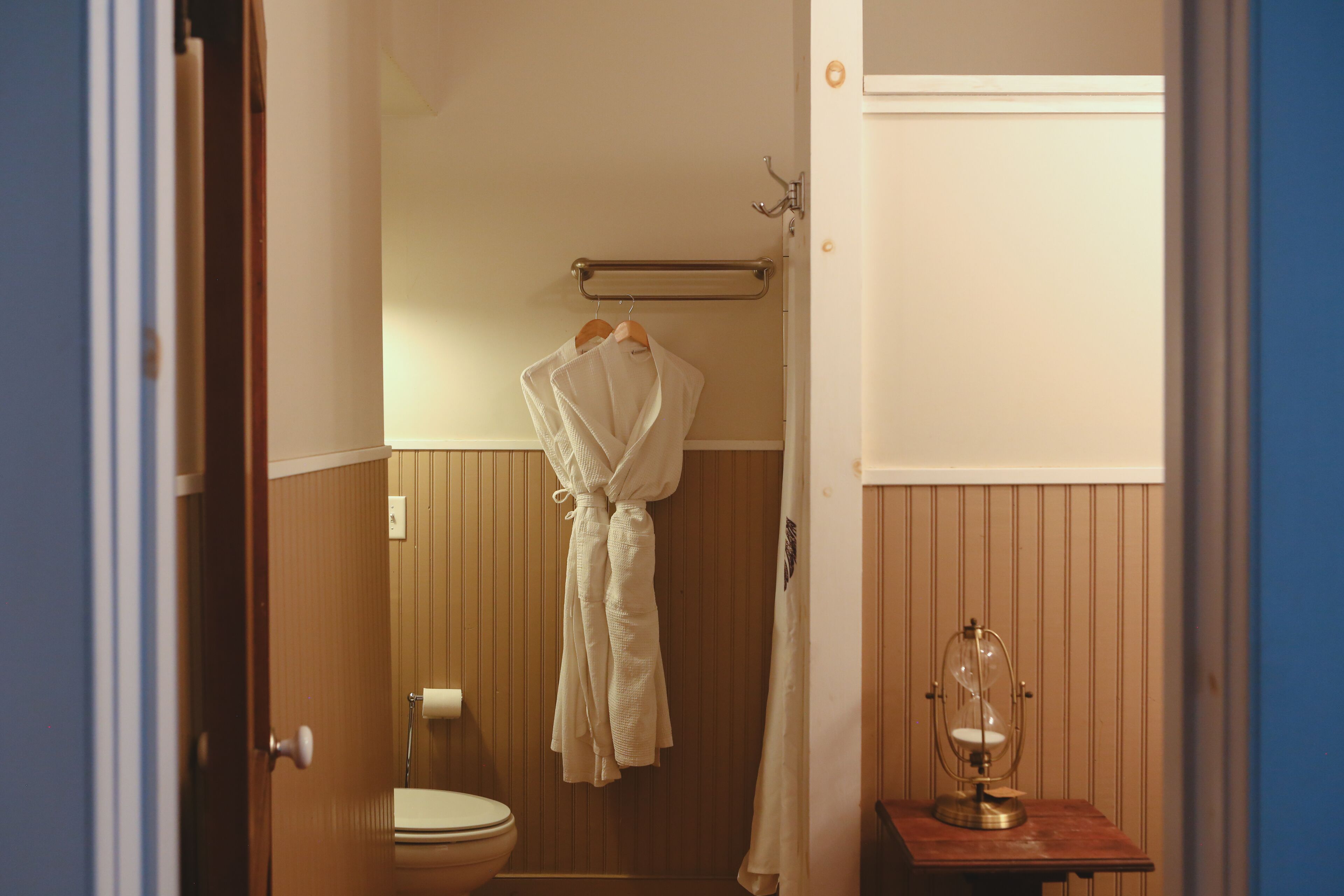 Une salle de bain comprenant des toilettes, un porte-serviettes avec deux peignoirs blancs, une commode en bois avec un sablier en verre, et un lambris en bois sur les murs.
