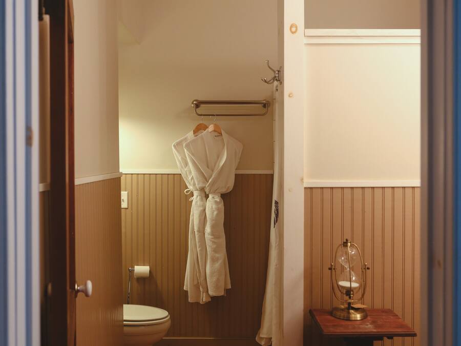 A bathroom with a toilet, a towel rack with two white robes, a wooden vanity with a glass hourglass, and wooden paneling on the walls.