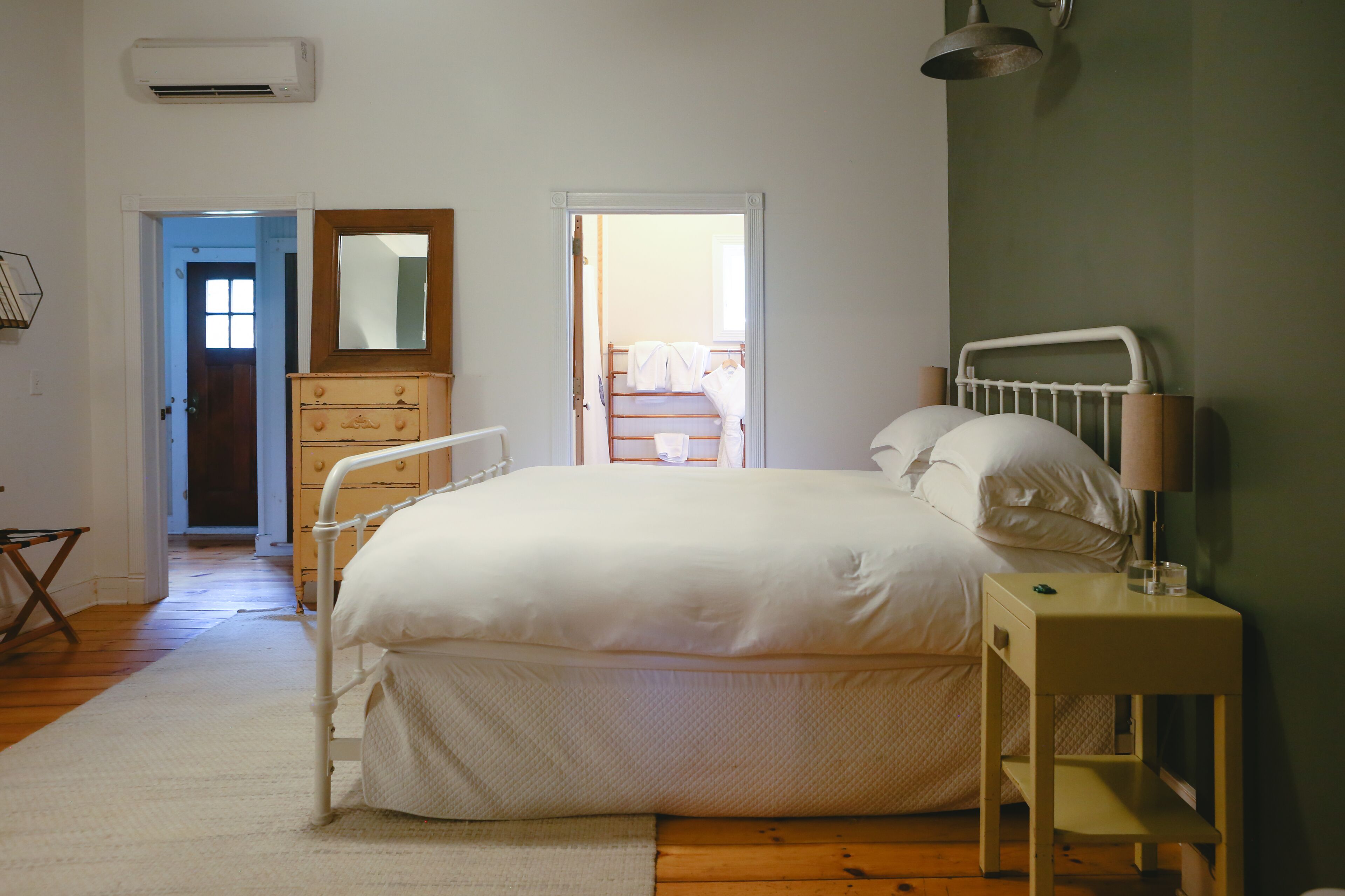 A bedroom with a bed, bedside table, and a dresser. There is a door leading to another room and a mirror above the dresser.
