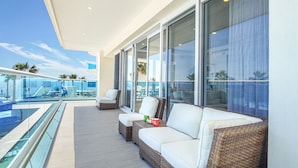 A balcony with a white sofa, wicker ottoman, and a glass railing overlooking a pool and palm trees.
