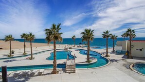 A pool area with multiple swimming pools, palm trees, and a beach in the background.
