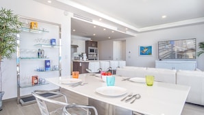 A modern kitchen with white cabinets, a dining area with a white table set for four, and a living area with a sofa and a painting on the wall.