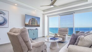 A spacious living room with a large window offering a view of the ocean, a ceiling fan, and a flat-screen TV mounted on the wall.