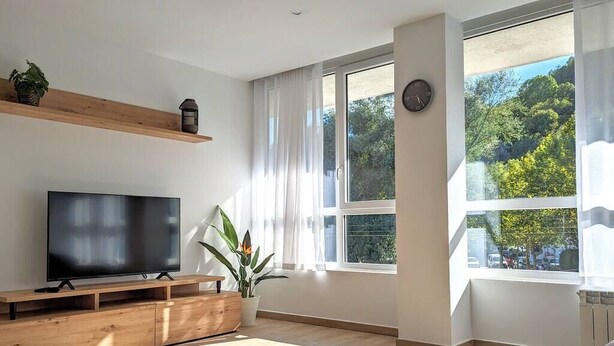 A modern living room with a flat-screen TV on a wooden stand, large windows with sheer curtains, and a view of greenery outside. - Hydros Freser Ripoll (Ripoll)