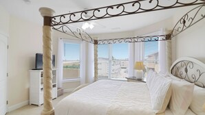 A bedroom with a four-poster bed, a television on a stand, and a view of buildings through large windows. - Oceanfront Condo with Pool & Stunning Beach Views (Tybee Island)