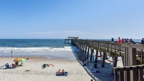 A beach with a wooden pier extending into the ocean, people relaxing on the sand, and a covered area at the end of the pier. - Oceanfront Condo with Pool & Stunning Beach Views (Tybee Island)