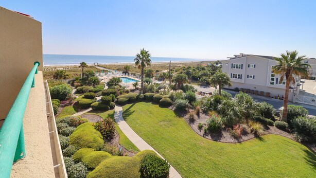 A view of a well-maintained garden with a swimming pool, palm trees, and a building with multiple windows. - Oceanfront Condo with Pool & Stunning Beach Views (Tybee Island)