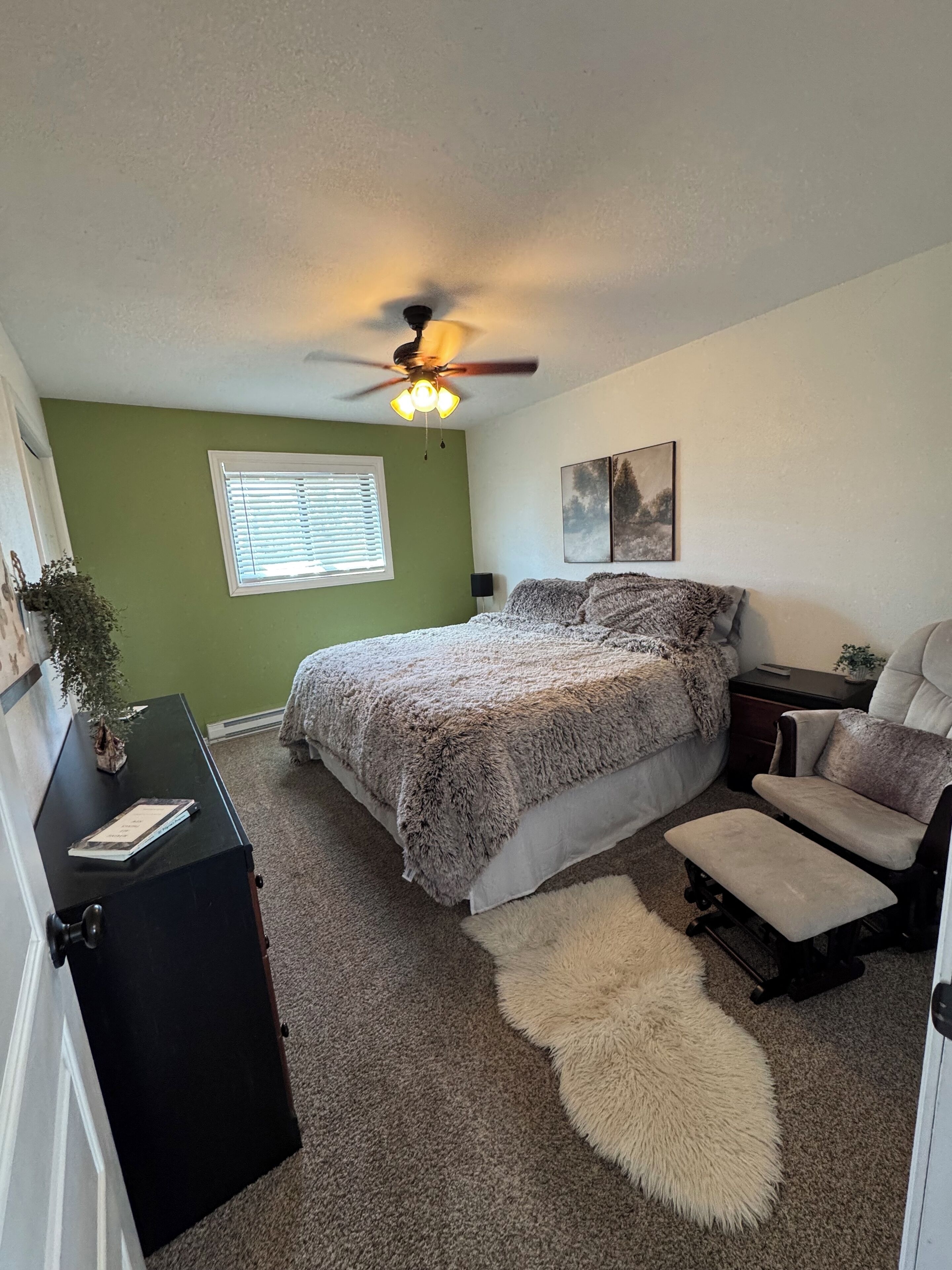 A bedroom with a bed, a desk, a chair, a rug, a ceiling fan, and a window with blinds.