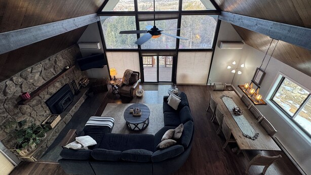 Un loft spacieux avec de hauts plafonds, une cheminée en pierre, un grand canapé sombre, une table à manger en bois et de grandes fenêtres offrant une vue sur le paysage.