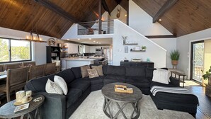 Un vaste espace de vie comprenant un grand canapé d’angle, une table basse ronde et une table à manger avec des chaises. On aperçoit une cuisine au fond de la pièce, et un escalier mène à l’étage supérieur.