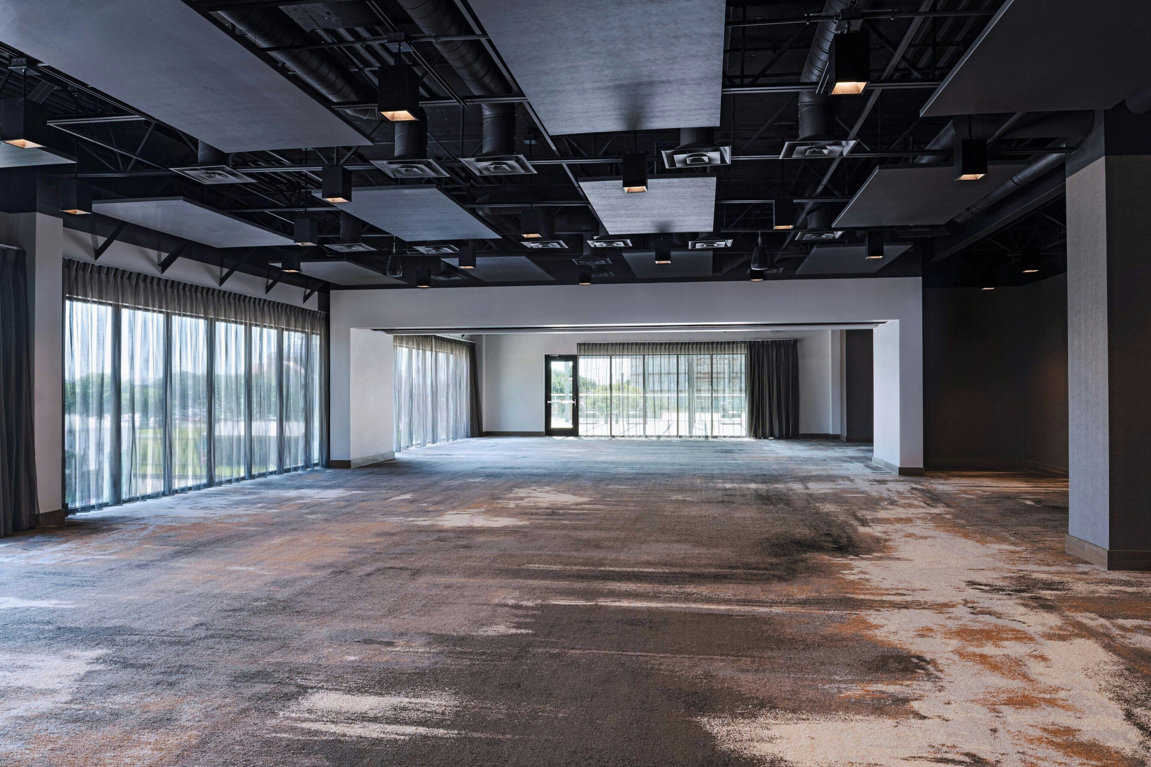 A spacious, industrial-style room with exposed ceilings, large windows, and a worn carpet.