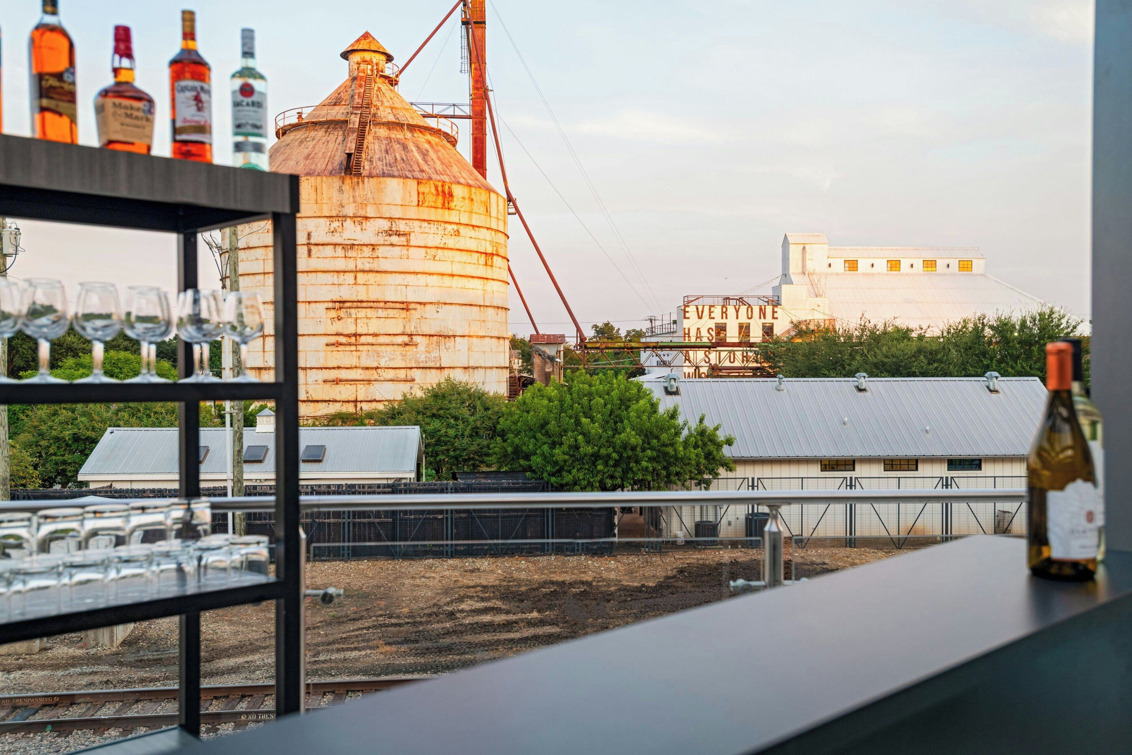 A rustic bar area with a large grain silo and industrial buildings in the background.
