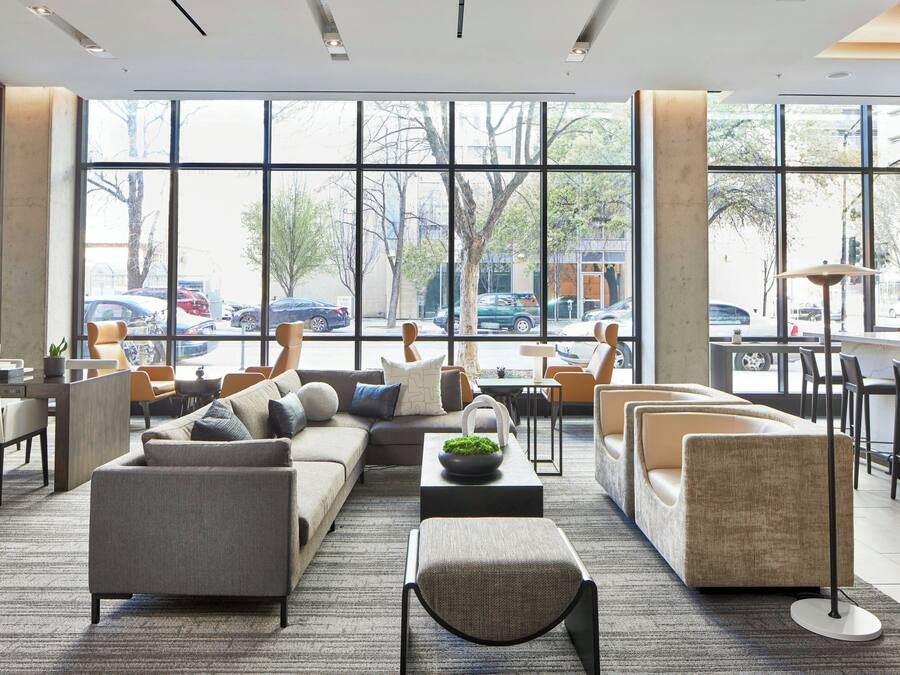 A modern lobby with a sectional sofa, armchairs, and a coffee table. Large windows offer a view of parked cars and trees outside.