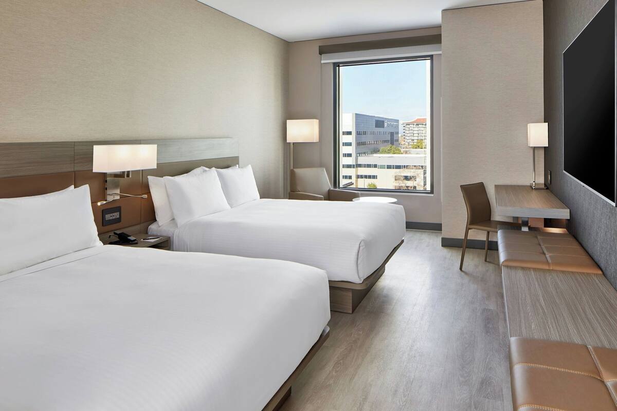 Hotelkamer met twee bedden, een bureau, een stoel en een groot raam met uitzicht op de stad.