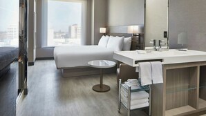 Una habitación de hotel moderna con una cama grande, una mesa redonda, un baño con tocador y espejo, y vistas de la ciudad a través de la ventana.