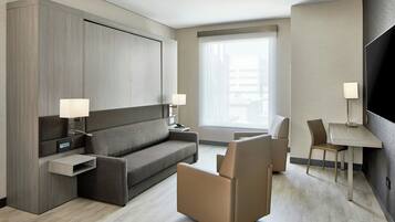 A modern hotel room with a grey sofa, a small table, a chair, and a TV.