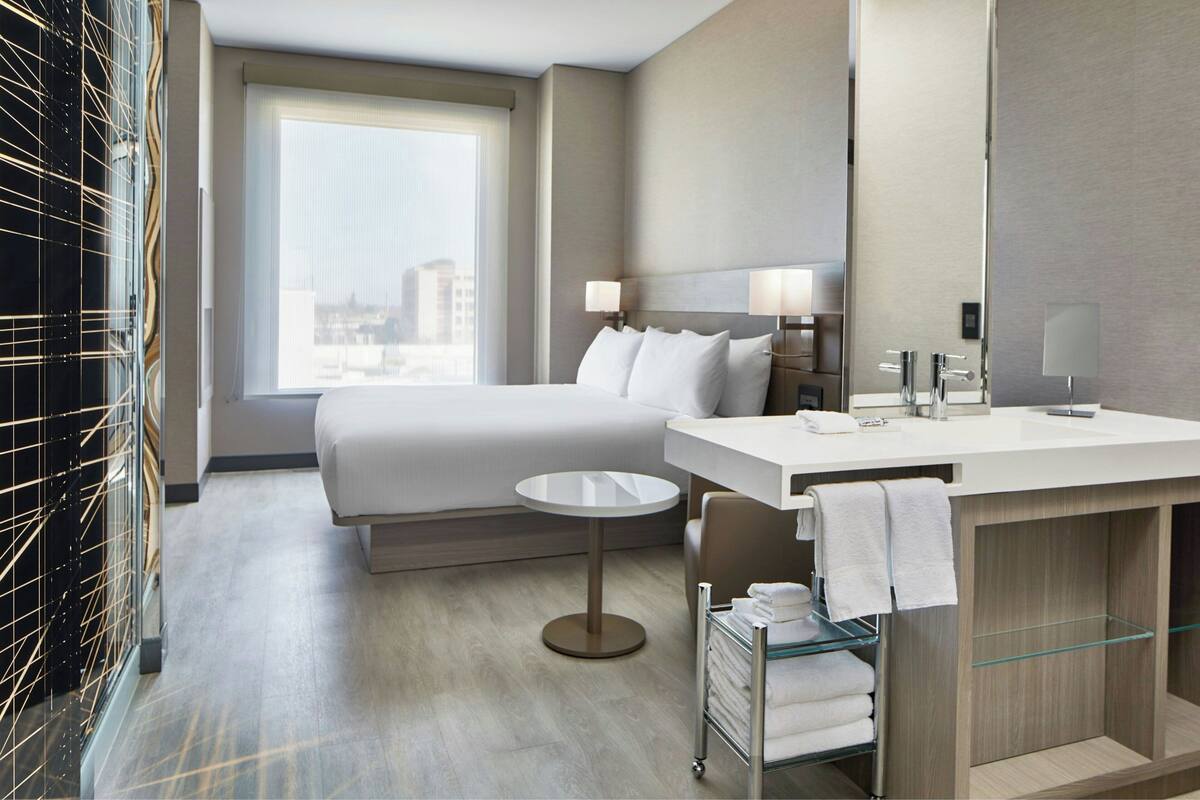 Hotelkamer met een bed, een rond tafel, een badkamer met wastafel en spiegel, en uitzicht op de stad.