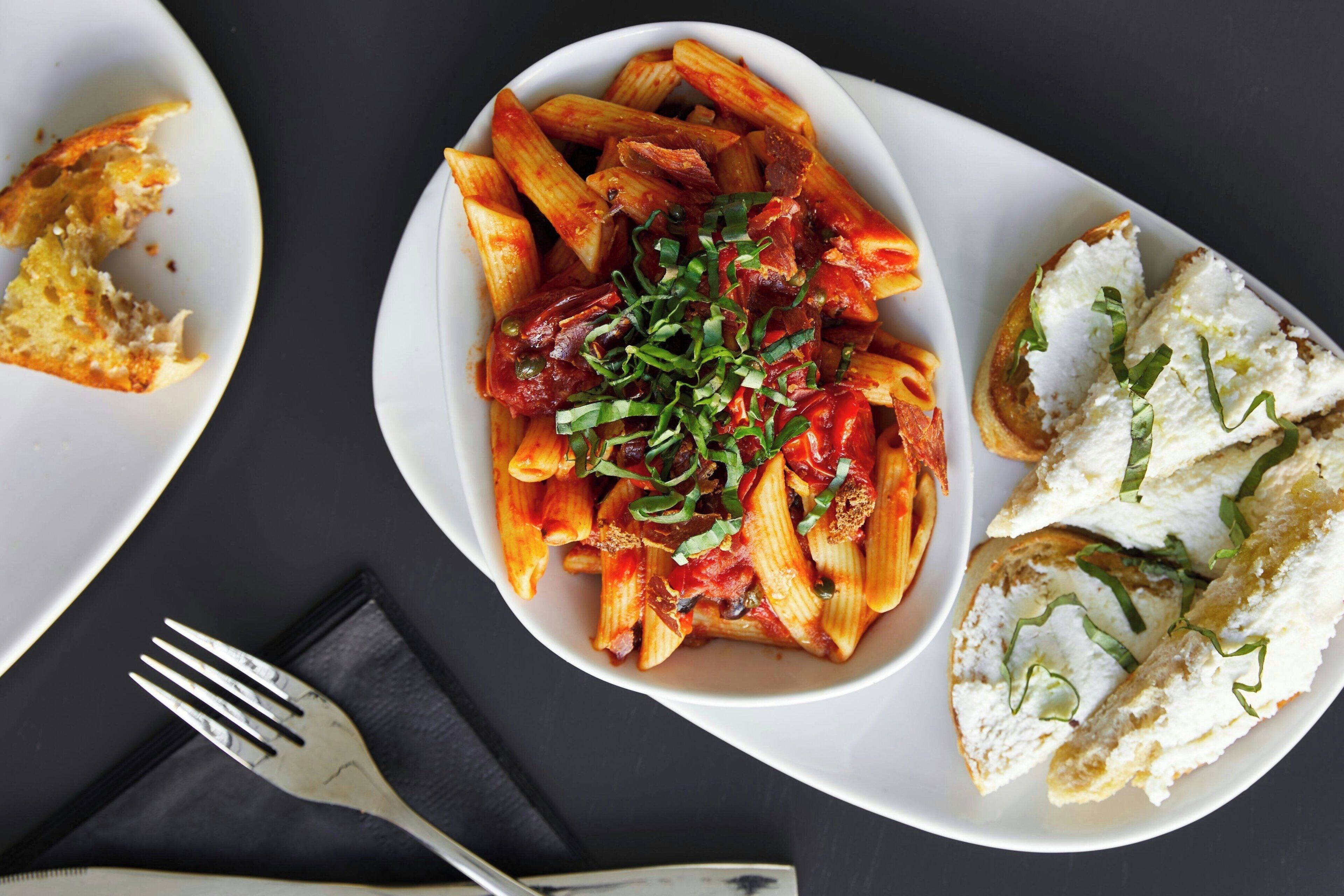 Een bord penne pasta met tomatensaus en groene uien, vergezeld van sneetjes brood.