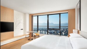 A modern hotel room with a large bed, a TV mounted on the wall, a chair, and a large window offering a city view.