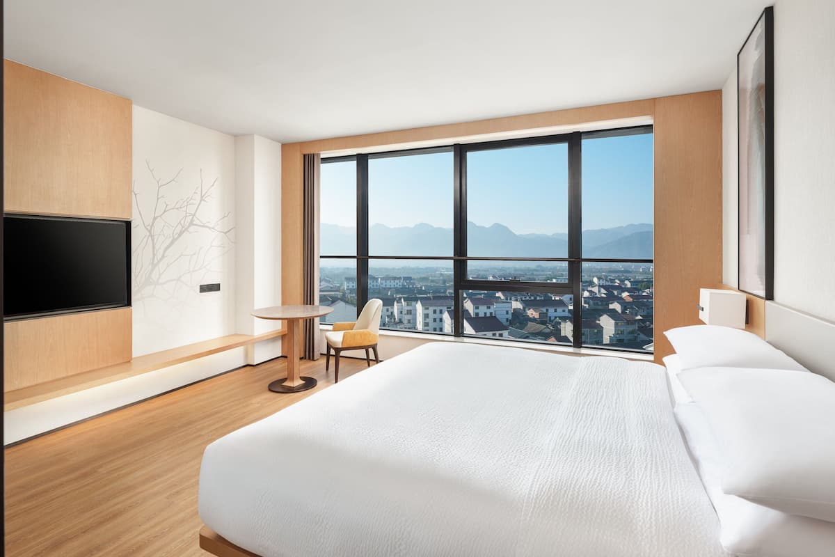 A modern hotel room with a large bed, a TV mounted on the wall, a chair, and a large window offering a city view.
