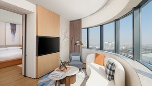 A modern hotel room with a curved wall, a flat-screen TV, a sofa, a chair, and a large window offering a city view.