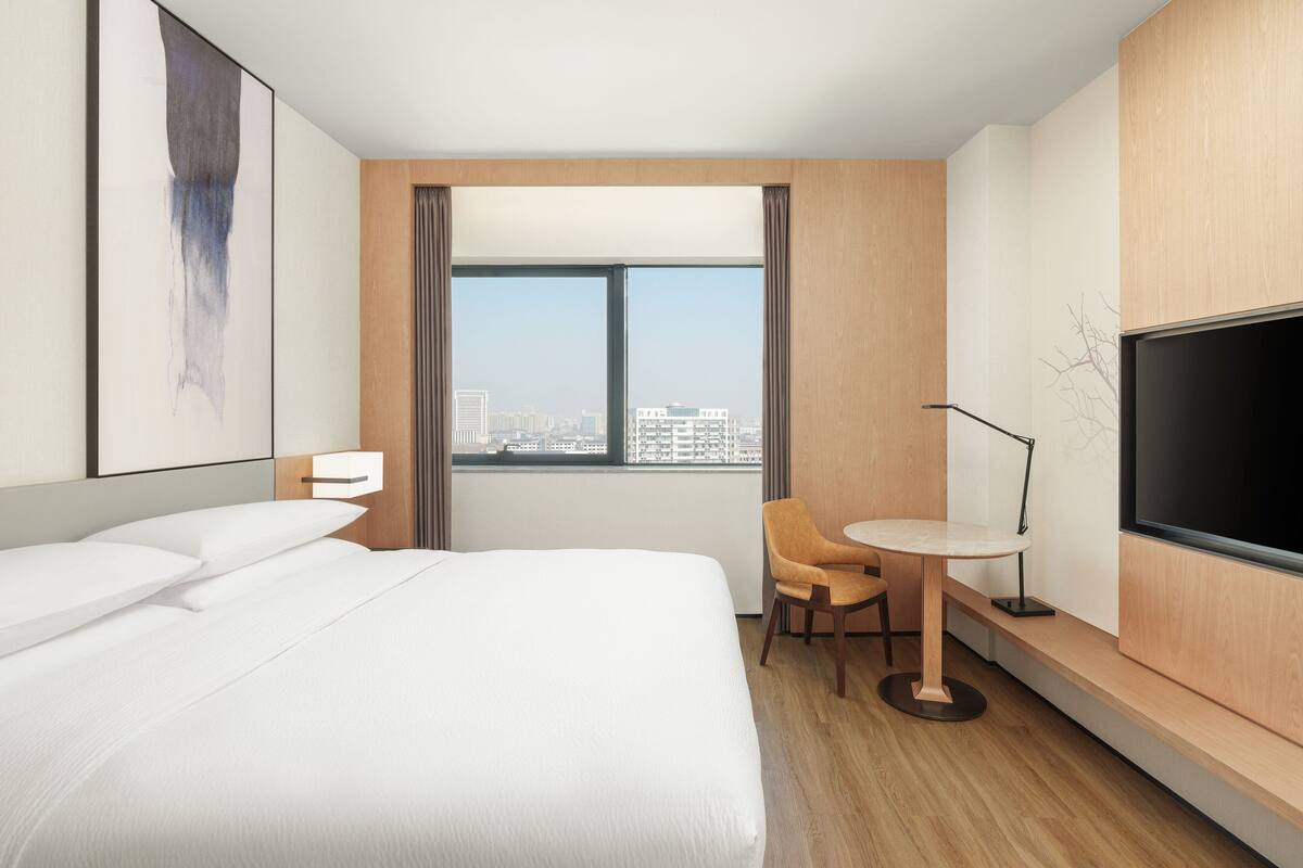 A hotel room with a large bed, a desk, a chair, a TV, and a window with a city view.