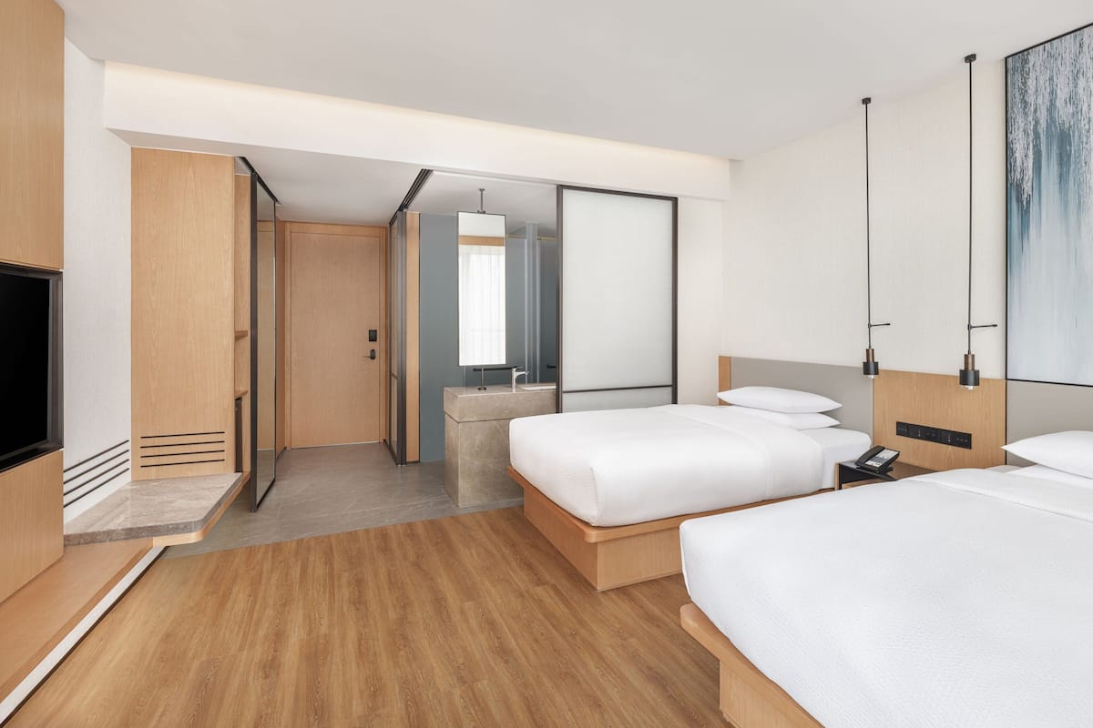 A modern hotel room with two beds, a large mirror, a vanity with a sink, and a flat-screen TV.