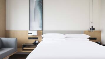 Una habitación de hotel moderna con una cama grande, una mesita de noche con lámpara, un escritorio pequeño y un sofá gris.