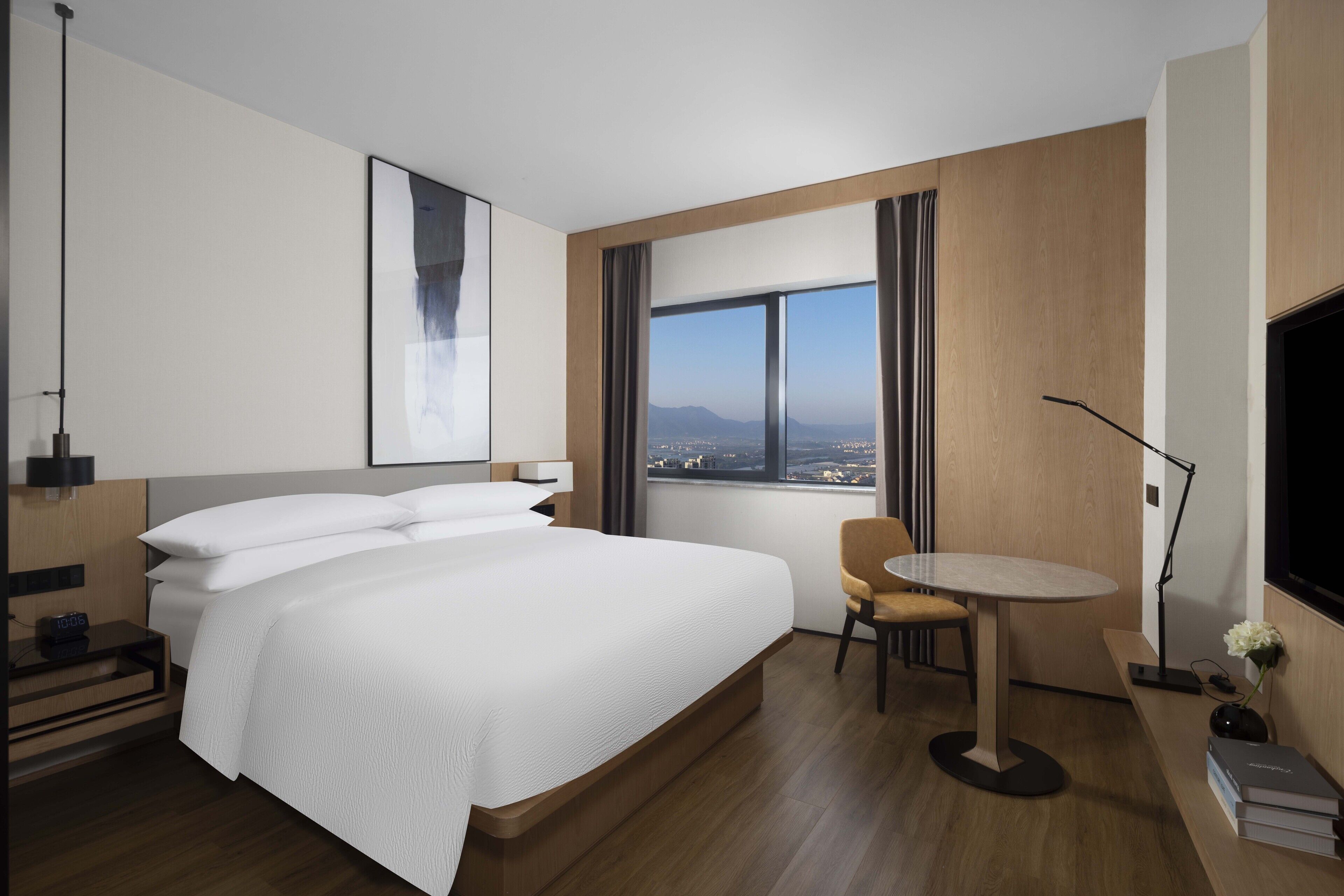 A modern hotel room with a large bed, a small round table, a chair, a desk, a TV, and a window with a city view.
